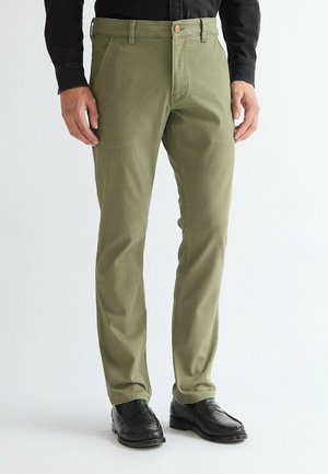 Man wearing olive green pants, black shirt, and black dress shoes standing against white background.