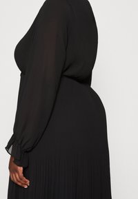 Robe noire plissée avec des manches longues en voile transparent et des poignets élastiques. Texture lisse avec des lignes verticales et une silhouette fluide.