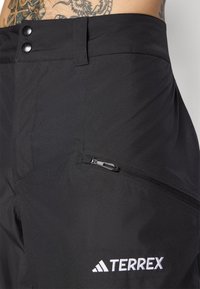 Pantalones exteriores negros con una textura suave, que cuentan con dos botones frontales, un bolsillo con cremallera y el logo blanco "TERREX" en el costado.
