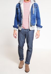 Hombre con chaqueta de mezclilla azul sobre sudadera gris, camiseta de rayas rojas y blancas, pantalones azul oscuro y botas de ante marrón, de pie sobre un fondo blanco.