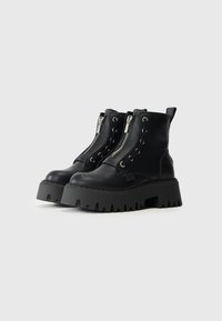 Botas de tobillo de cuero negro grueso con cremalleras plateadas en la parte delantera, suelas gruesas y estriadas, y tiradores en un fondo blanco.