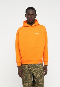Felpa arancione con tasca frontale, dotata di un piccolo logo bianco. Indossata con pantaloni a motivo mimetico, evidenziando una vestibilità rilassata.
