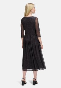 Langes, marineblaues Kleid mit durchsichtigen langen Ärmeln, floralem Muster, gerafftem Taillenbereich und einem knöchellangen Saum; kombiniert mit schwarzen Blockabsatzsandalen.