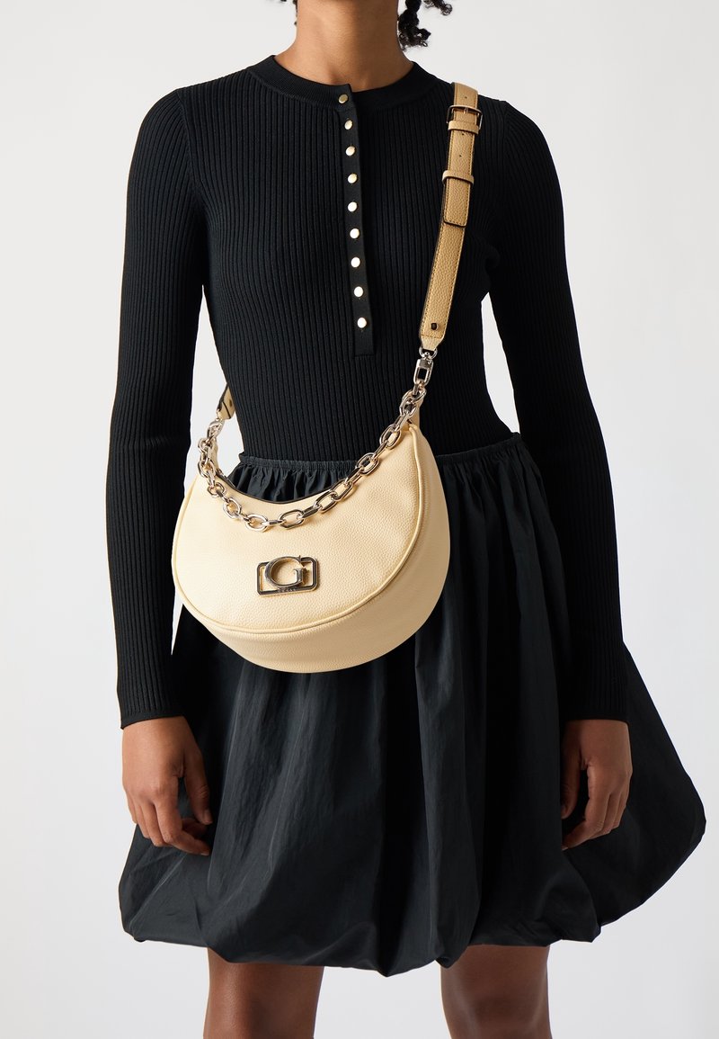 Femme portant un haut côtelé noir à manches longues et une jupe bouffante avec un sac bandoulière beige en forme de croissant, orné d’une chaîne et d’un fermoir.