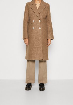 Kvinde iført en knælængde, dobbeltradet brun tekstureret frakke, beige bukser med vide ben og sorte snørestøvler, stående mod en hvid baggrund.