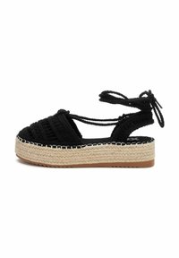 Sandal de esparto tejido negro con una gruesa plataforma de yute y lazos en el tobillo. Presenta una parte superior texturizada y costuras blancas en contraste a lo largo de la suela.