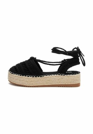 Sandal de esparto tejido negro con una gruesa plataforma de yute y lazos en el tobillo. Presenta una parte superior texturizada y costuras blancas en contraste a lo largo de la suela.