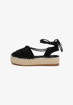 Sandal de esparto tejido negro con una gruesa plataforma de yute y lazos en el tobillo. Presenta una parte superior texturizada y costuras blancas en contraste a lo largo de la suela.