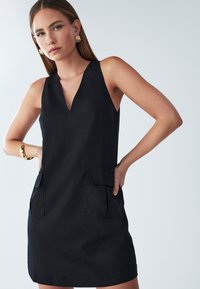 Ženska s ravnimi lasmi, ki nosi zlate uhane in zapestnico, oblečena v črno obleko brez rokavov s sprednjimi žepi, pozira z rokama na bokih.