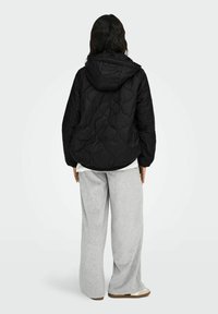 Schwarze gesteppte Jacke mit Kapuze, die ein strukturiertes Muster aufweist. Kombiniert mit lockeren grauen Jogginghosen. Ansicht von hinten.