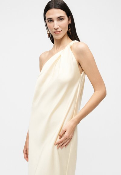 Γυναίκα με μακριά σκούρα μαλλιά φορώντας κρεμ φόρεμα με έναν ώμο και καφέ κρεμαστά σκουλαρίκια, πόζαρε μπροστά σε απλό ανοιχτό φόντο.