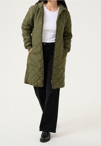 Quilted olive green long coat with a hood, featuring side pockets and front zipper. Worn over a white shirt and black pants.