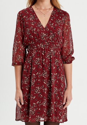 Robe florale rouge avec taille élastique, col en V et manches trois-quarts transparentes, portée avec des colliers délicats et des bagues aux deux mains.