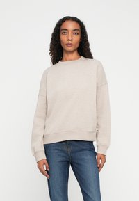 Mujer con cabello rizado de pie, vestida con un sudadera de manga larga beige y jeans azules, contra un fondo blanco liso.