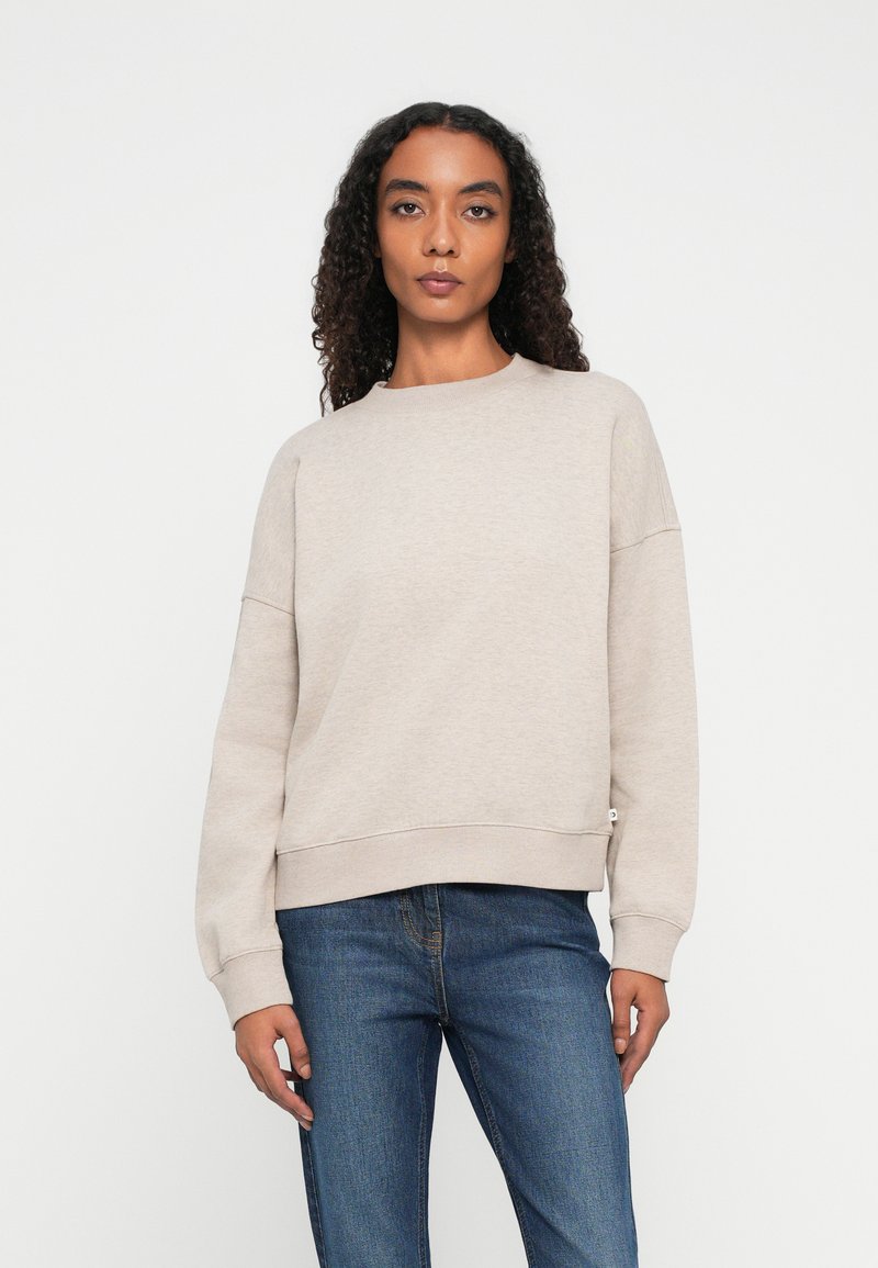 Mujer con cabello rizado de pie, vestida con un sudadera de manga larga beige y jeans azules, contra un fondo blanco liso.