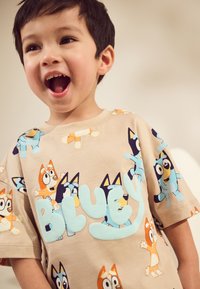 T-shirt beige avec des chiens dessinés de manière ludique en cartoon, de différentes couleurs et postures, accompagné d'un texte bleu "BLUEY" en appliqué doux et proéminent.