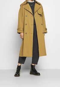 Khaki trench coat s dvouřadým designem, velkými klopami a knoflíkovými detaily. Nosit over tmavě šedým topem a ladícími zúženými kalhotami a černými botami.
