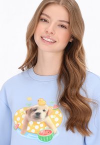 Világoskék pulóver, amelyen egy grafika látható: egy kölyökkutya sárga úszógumiban, mellett egy gumimadár, körülöttük görögdinnye szeletek és napos háttér.