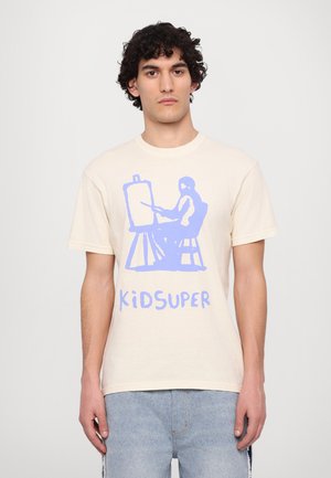 Camiseta de algodón crema con un gráfico artístico azul de una figura pintando en un caballete, con el texto "KIDSUPER" en la parte inferior.