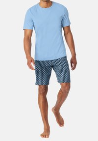 Světle modré tričko s kulatým výstřihem, doplněné tmavě modrými plavkami s potiskem ve geometrických tvarech. Bez obuvi.