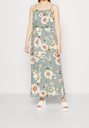 Maxi-jurk van lichtgroene stof met bloemenprint in oranje en wit. Heeft dunne bandjes en een elastische taille. Draagt witte blokhakken.