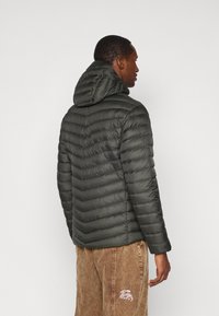 Muškarac u tamnozelenoj qiltanoj jakni s kapuljačom i smešnim kordonac hlačama s bijelim grafičkim motivom na džepu, okrenut bočno prema bijeloj pozadini.