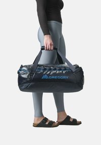 Gregory duffel bag, navy with blue accents, textured nylon, zippered closure, padded handles; held by a person in gray leggings and sandals.