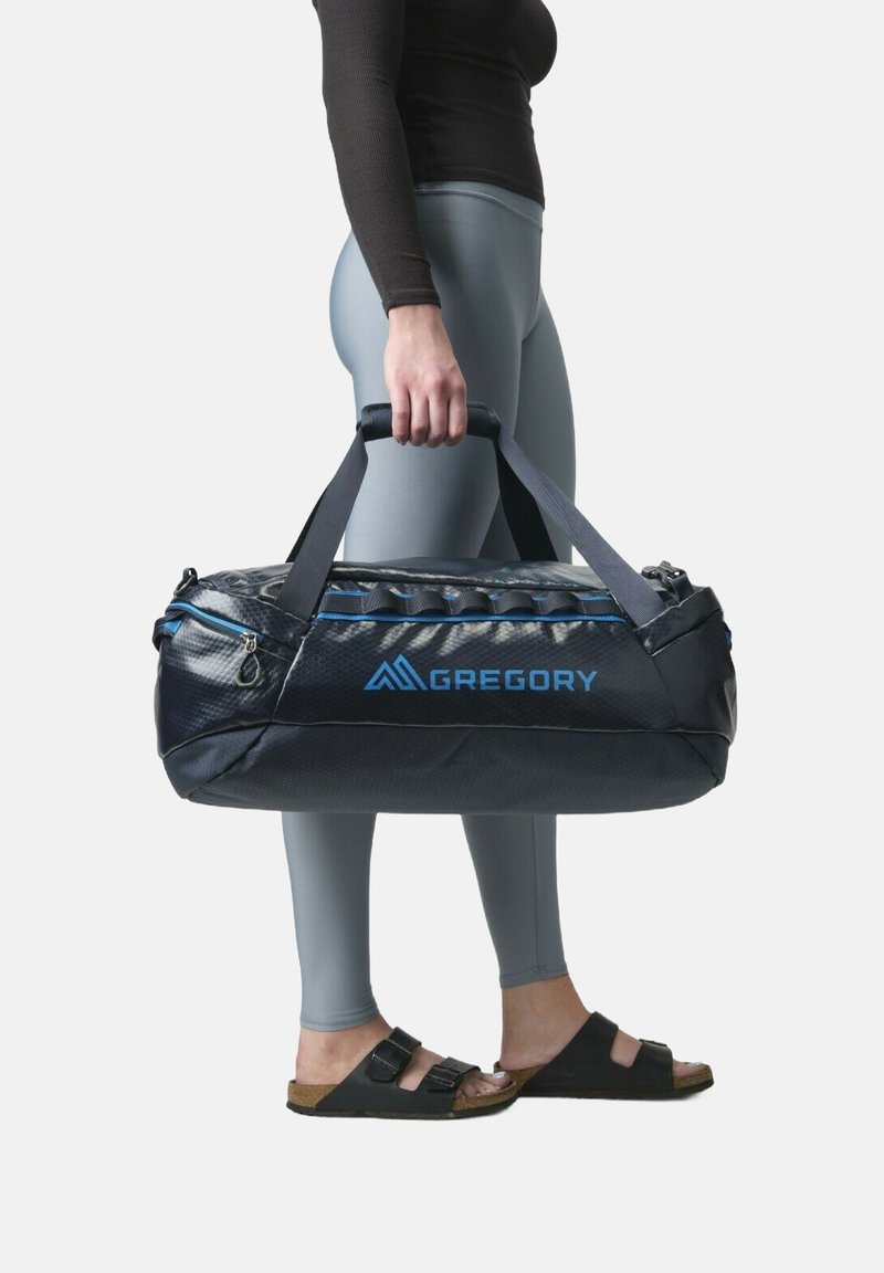 Gregory duffel bag, navy with blue accents, textured nylon, zippered closure, padded handles; held by a person in gray leggings and sandals.