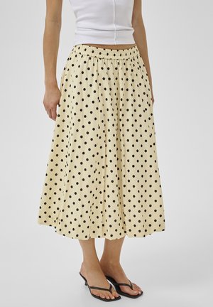 Žena nosi krémovú midi sukňu s čiernymi bodkami, kombinovanú s bielym tielkom bez rukávov a čiernymi sandálmi na podpätku.