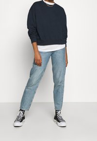 Felpa blu navy con maniche lunghe e orlo a costine, abbinata a jeans dritti azzurri. Sneakers nere con suole bianche completano l'outfit.