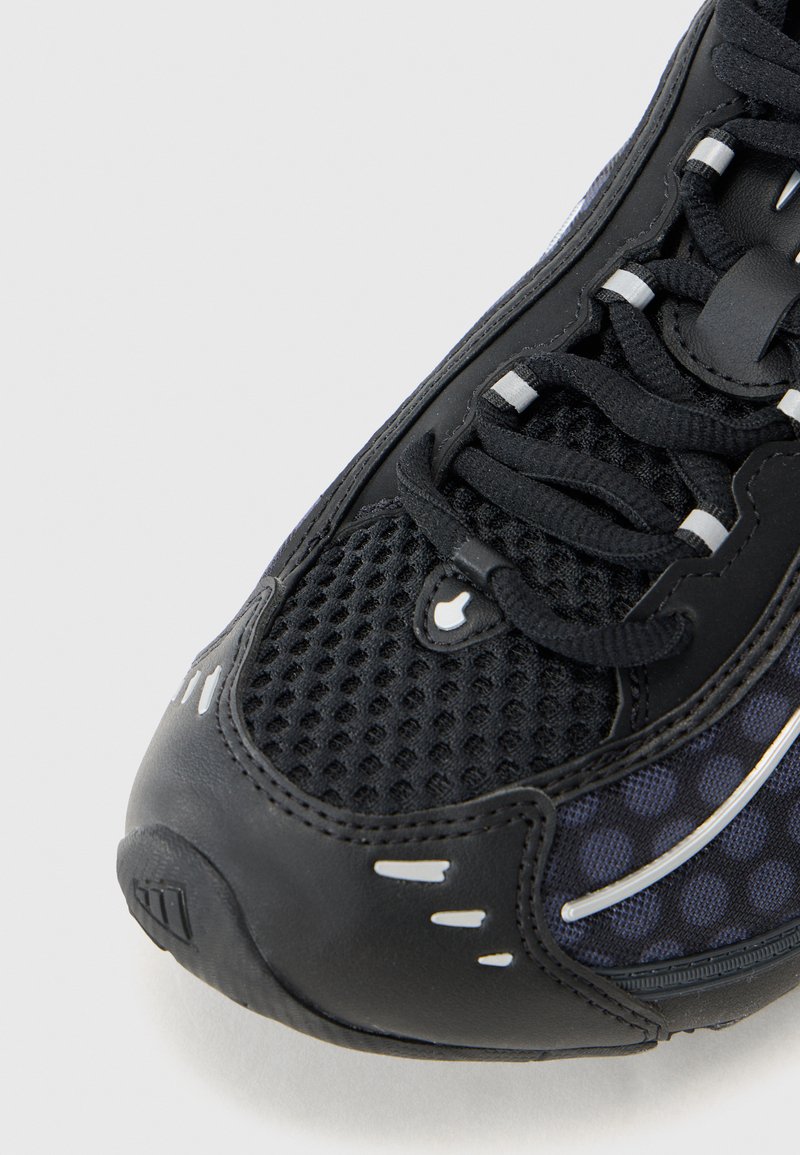 Zapatilla deportiva negra con parte superior de malla texturizada, puntera redondeada y acentos en blanco contrastantes a lo largo del costado y los cordones.