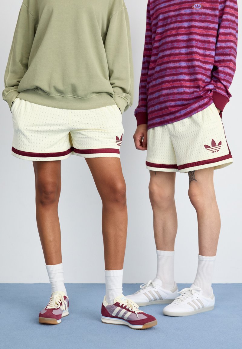 Shorts blancs avec des accents bordeaux et logo Adidas ; tissu texturé. Sweat-shirt gris-vert ; chemise à manches longues rayée violette. Chaussures décontractées.