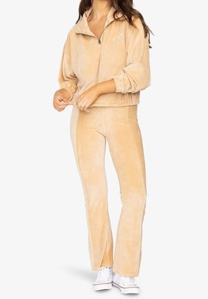 Vrouw draagt beige corduroy half-rits trui en bijpassende uitlopende broek, gecombineerd met witte sneakers, staand op een witte achtergrond.