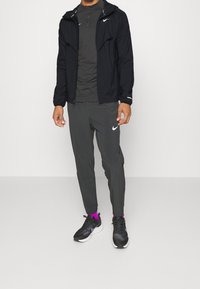 Mężczyzna ubrany w czarną kurtkę Nike, ciemnoszary rozpinany sweter, ciemnoszare dresy z logo Nike, czarne trampki i jasnofioletowe skarpetki stojący.