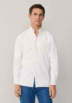 Uomo che indossa una camicia bianca con bottoni e un logo azzurro chiaro sul petto, e jeans blu, in piedi contro uno sfondo grigio chiaro uniforme.