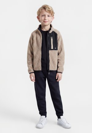 Jeune garçon blond debout face à l'avant portant une veste polaire beige avec des accents noirs, pantalon noir et baskets blanches sur fond blanc.
