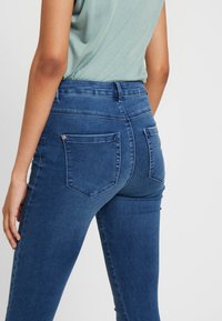 Calças skinny em denim azul escuro. Apresentam uma cintura alta, dois bolsos traseiros e costura subtil. Textura suave com um ligeiro elástico.