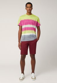 Tie-dye t-shirt i grön, rosa och grå med korta ärmar. Klockar med vinröda shorts. Accessoarer inkluderar vita sneakers med beige detaljer.