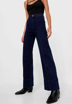 Vrouw draagt donkerblauwe, hooggetailleerde jeans met wijde pijpen, zwarte enkellaarsjes met puntige neus en een zwarte mouwloze top met dunne bandjes.