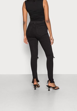 Vaqueros skinny de talle alto en negro con dobladillos desgastados, hechos de denim. Combinados con tacones abiertos negros, mostrando un diseño minimalista.