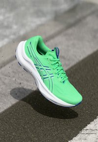 Neon-grüner Sportschuh mit strukturiertem Stoff, erhöhten blauen Akzenten und runden Perforationen. Leichte Sohle, die für Komfort sorgt.