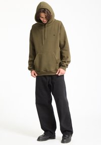 Olivengrønt hoodie med snøre i hætten og forlommer, parret med løstsiddende sorte bukser og sorte sko, stående foran en hvid baggrund.