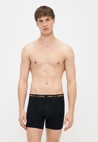 Černé boxerky s přiléhavým střihem, se žlutým pasem s logem, vyrobené z měkké bavlněné látky. Jednoduchý a minimalistický styl.