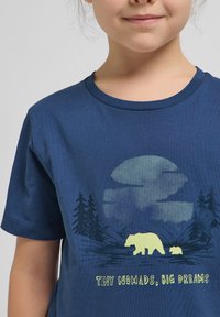 Tamsiai mėlynas medvilnės marškinėliai su lokio ir lokiuko grafika, apsupta medžių ir kalnų, su tekstu "MAŽIEJI NOMADAI, DIDELI DREAMAI."