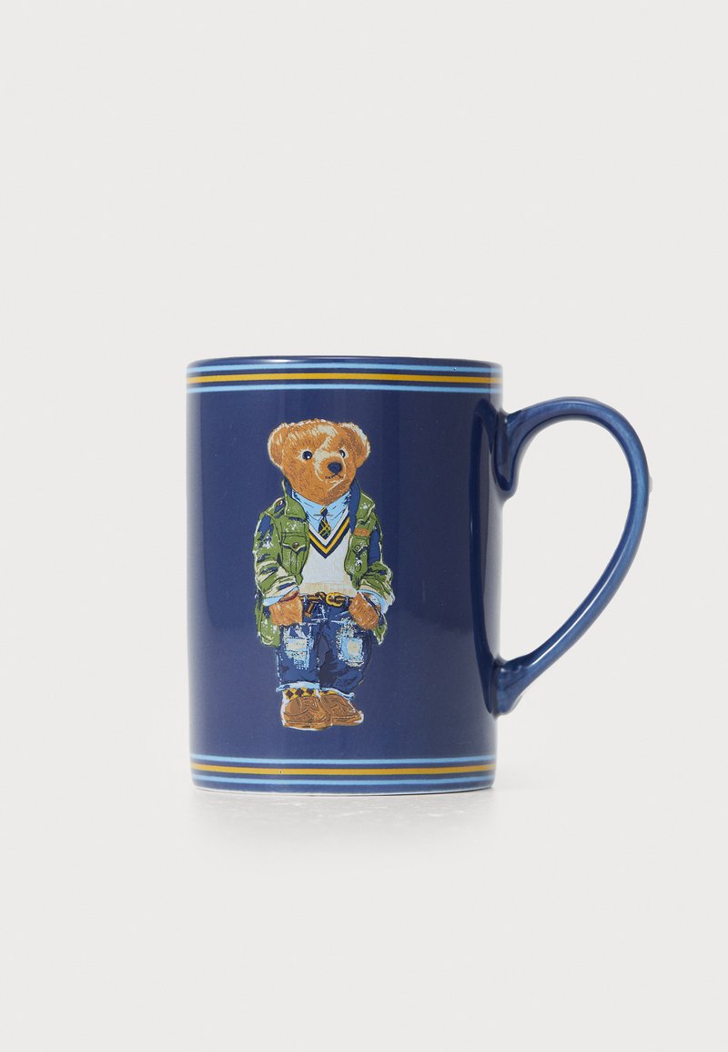 Blue ceramic mug featuring a bear illustration wearing a green jacket and denim. Gold and blue stripes decorate the rim. Tapered shape.
