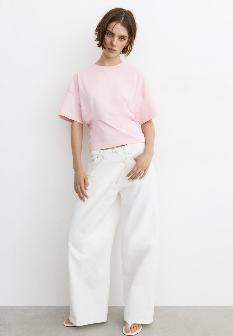 Jeune femme portant un haut rose ample à manches courtes et un pantalon blanc large, debout contre un mur blanc dans une posture détendue.