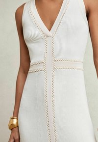 Rochie tricotată fără mâneci, de culoare bej deschis, cu decolteu adânc în V, textură cu nervuri și accente decorative împletite de-a lungul cusăturilor.