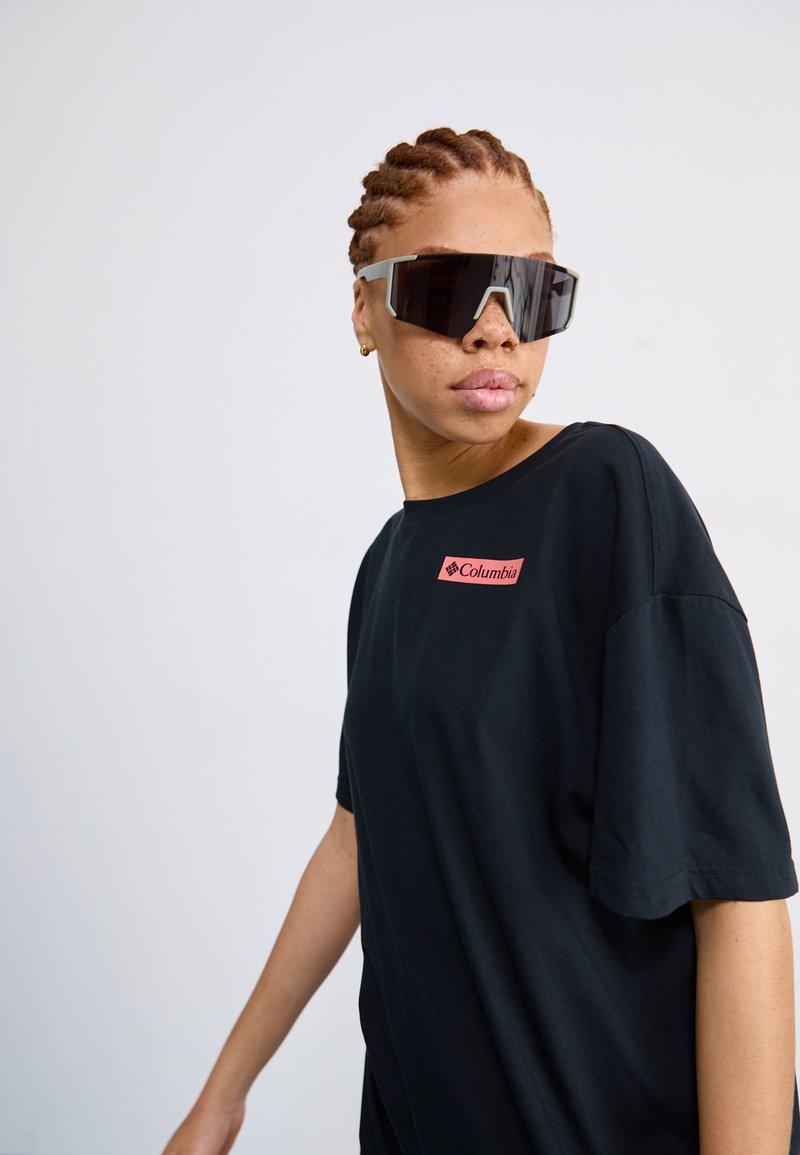 Camiseta negra de manga corta con un logo rojo de Columbia en el pecho. Modelo lleva gafas de sol negras de gran tamaño con un diseño geométrico.