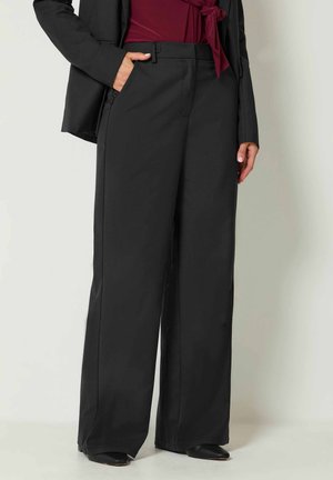 Femme portant un pantalon noir à jambes larges, un blazer noir et un chemisier bordeaux avec un détail noué, debout avec une main dans la poche sur un fond uni.