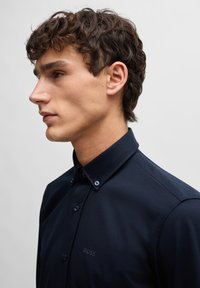 Marineblaues, knöpfbares Hemd mit strukturiertem Kragen, das dezente Branding auf der Brust aufweist und aus glattem, texturiertem Stoff besteht.